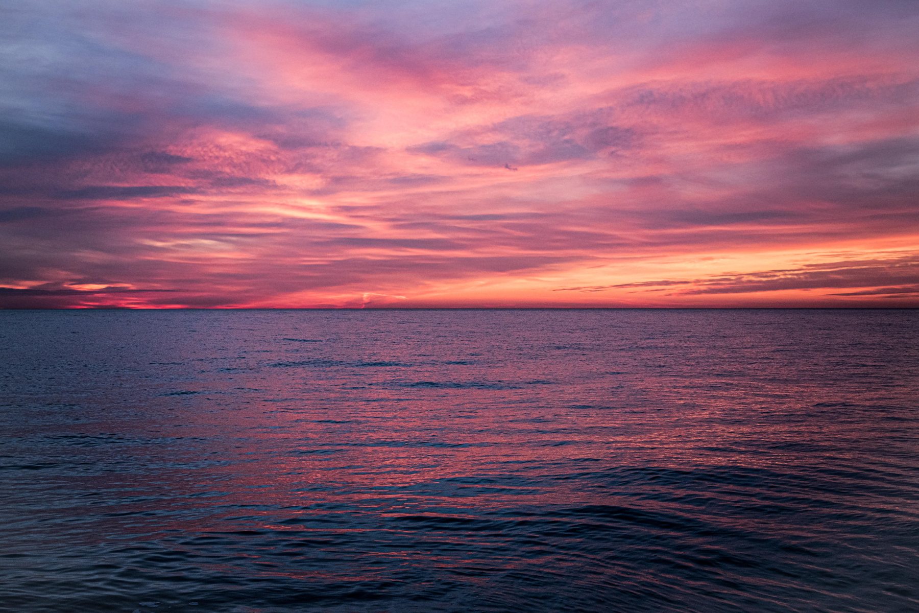 A vivid magenta and pink sunset over calm water, with a thin orange glow at the horizon and pink reflections on the dark water